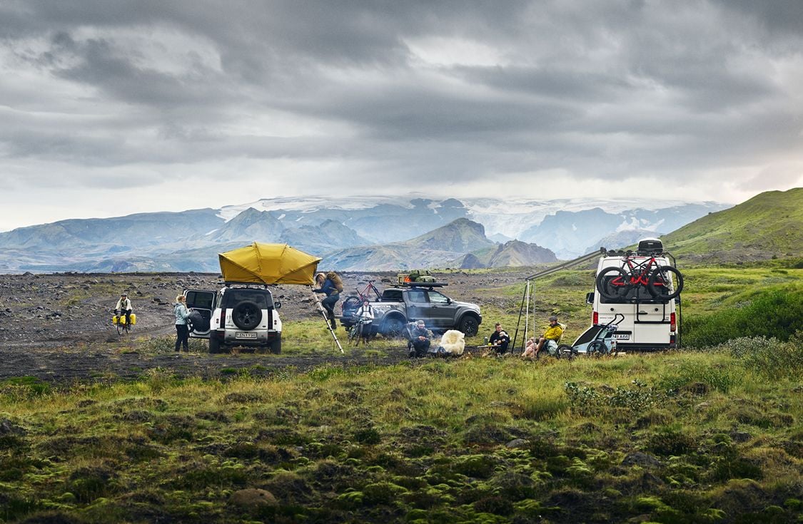 Outdoor campsite with vehicles equipped with Thule gear in a scenic mountain landscape. People are gathered around tents and camper vans with rooftop tents, bikes, and other outdoor equipment.