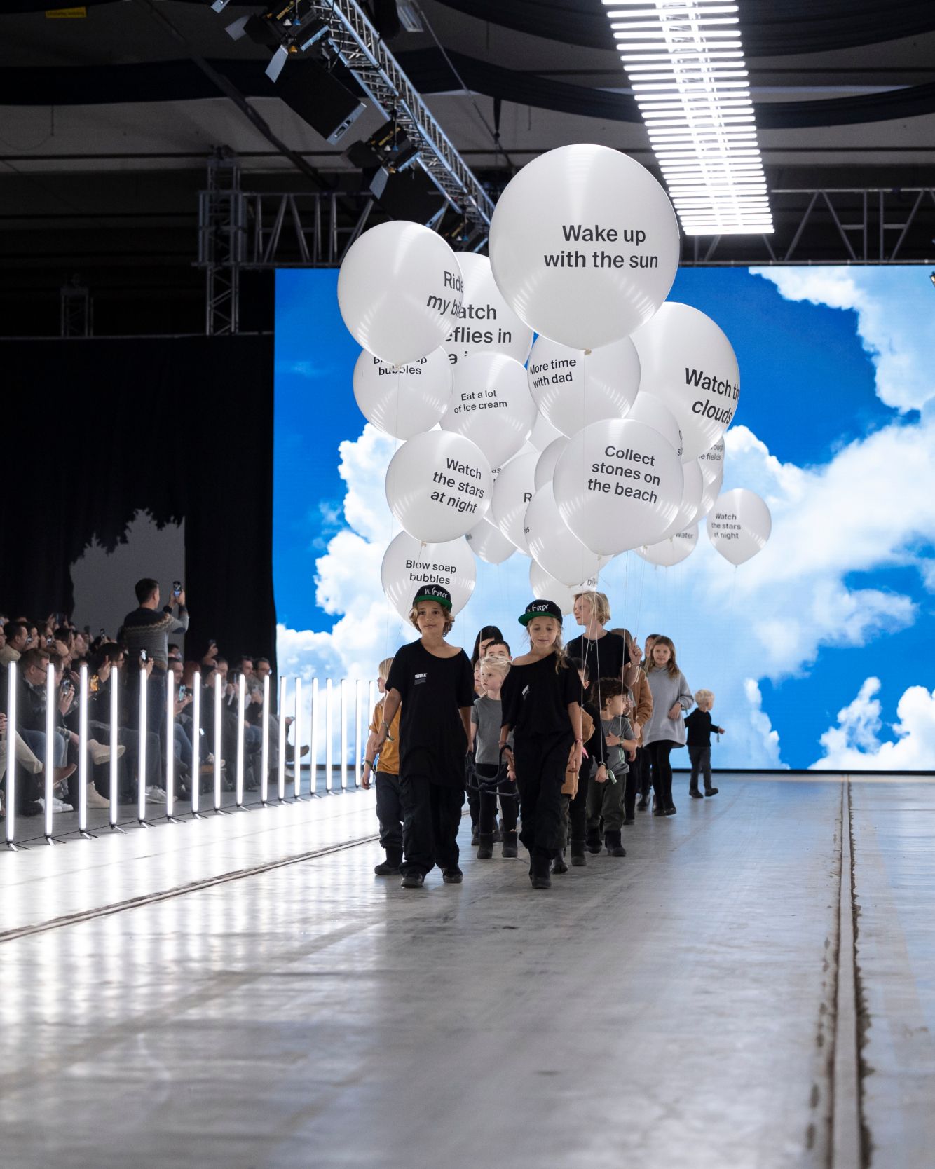A group of children and toddlers walking down a fashion runway holding big white balloons with text on them. Behind them is a big screen with a  blue sky and clouds and a group of people watch the screen.