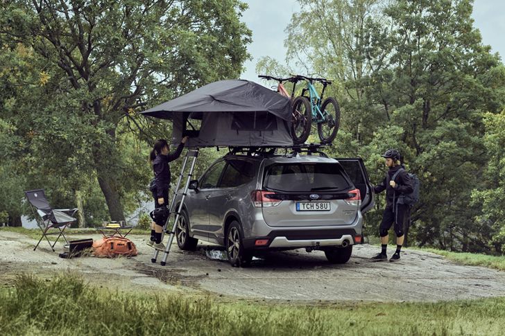 Alt text A woman climbs the ladder into a rooftop tent parked in the forest with bikes on the roof of the car.