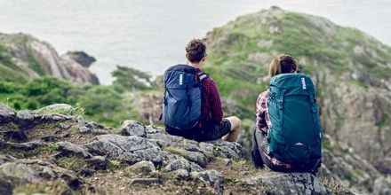 Two people sit on a rocky coast overlooking the ocean wearing a backpack for men and a backpack for women.