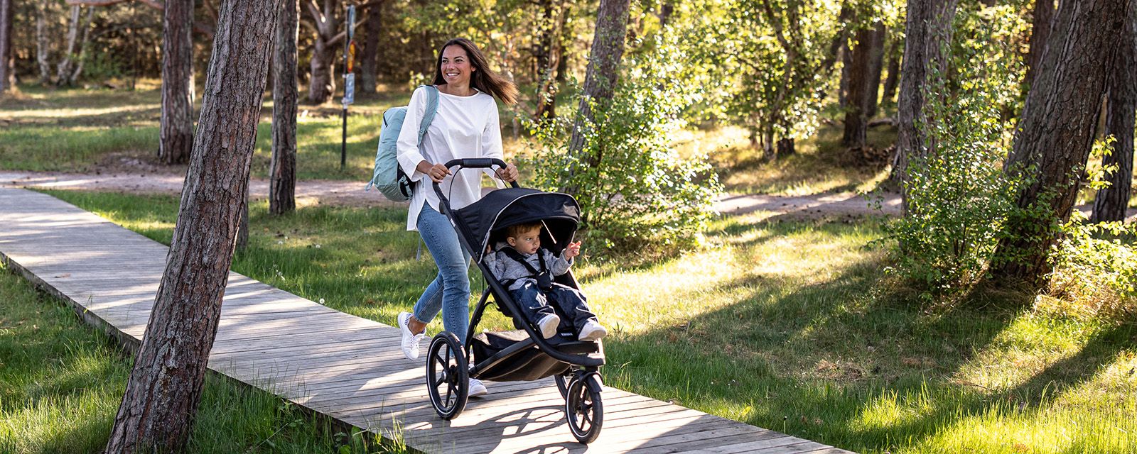 Eine Mutter und ihre zwei Kinder spazieren mit einem Thule Urban Glide 2 Jogging-Buggy für jeden Untergrund durch einen Park.