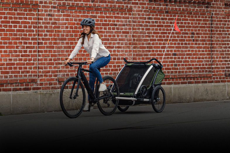 A woman bikes with her green Thule Chariot Cab bike trailer, there is a brick wall in the background.