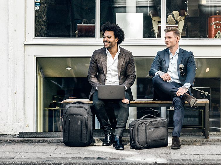 Two men with laptop bags and laptop sleeves sit outside a shop on a bench, one has his laptop open.