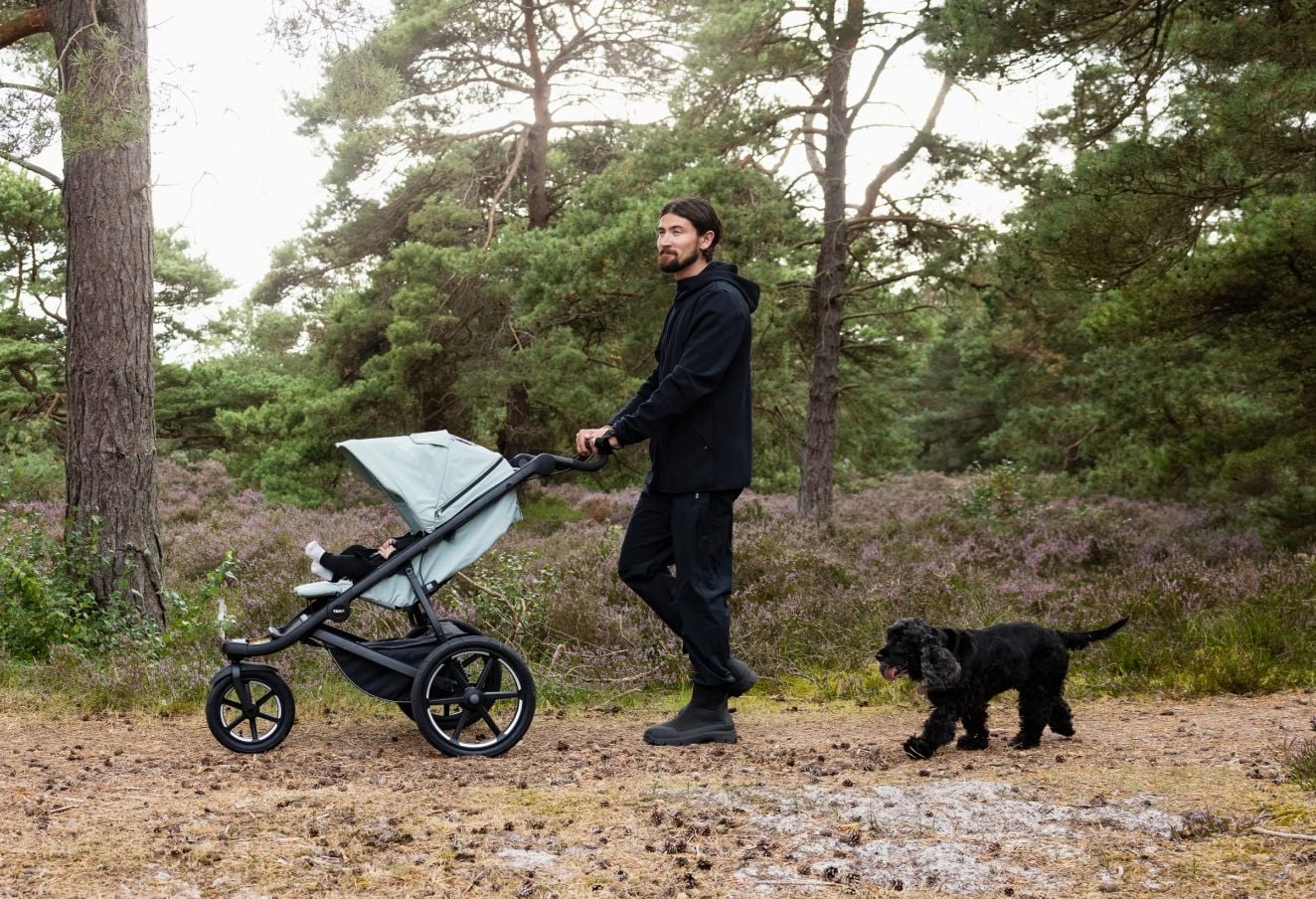 Un homme marchant avec une poussette Thulé sur un sentier forestier tandis qu’un chien marche à ses côtés.