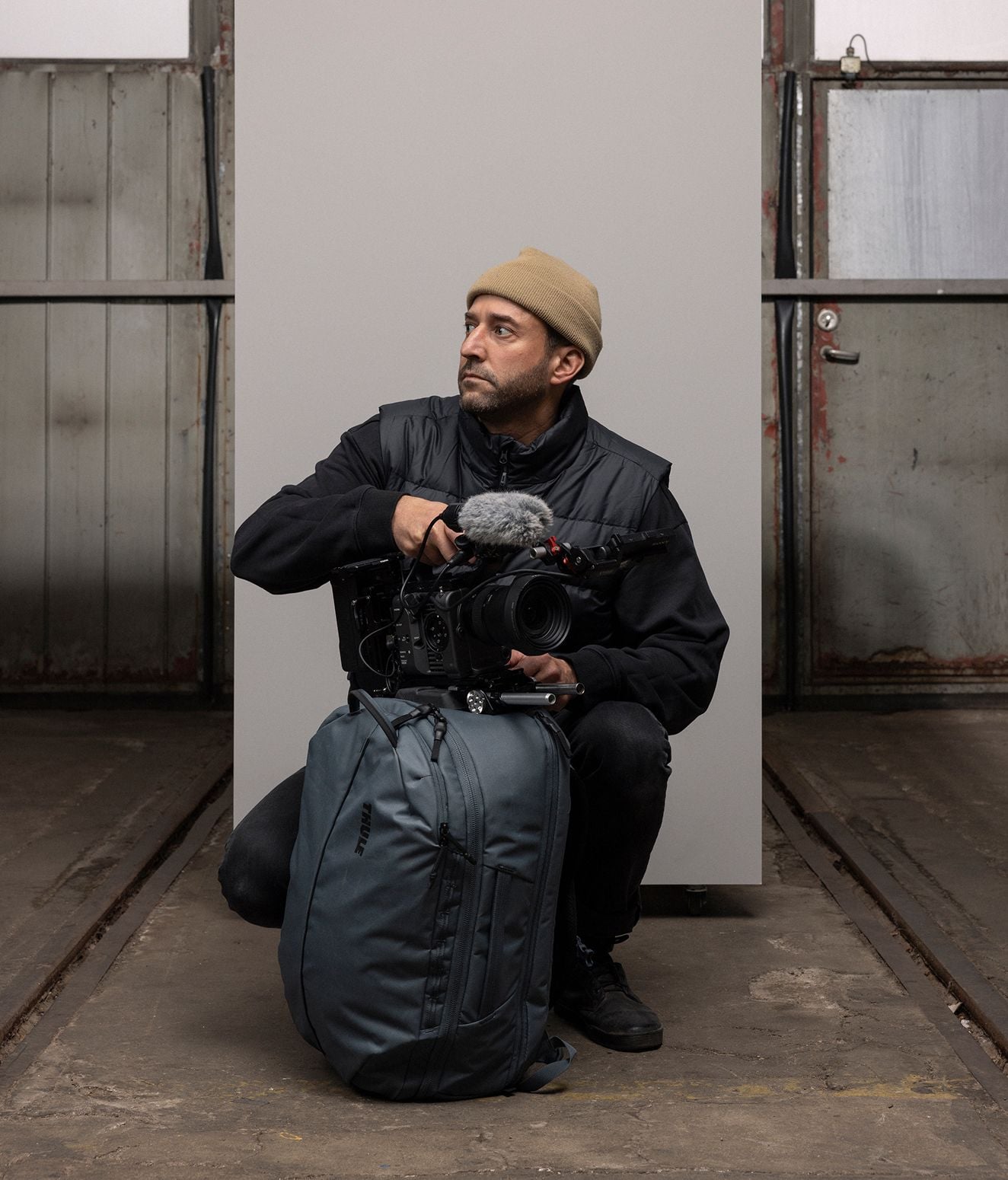 Eric Crosland hält eine professionelle Kamera in der Hand, während seine Hand auf einer Thule Aion Reisetasche ruht. Er trägt eine Mütze und eine schwarze Weste und schaut an der Kamera vorbei.