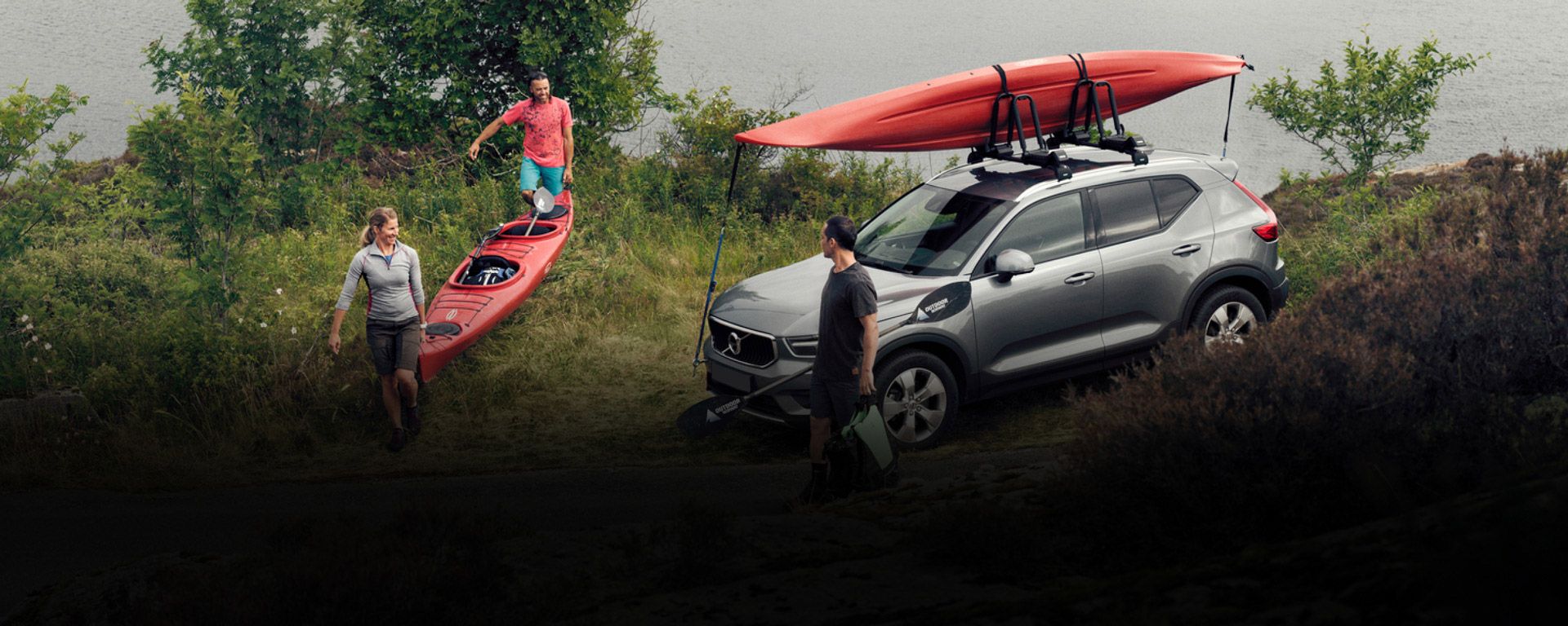 Two people carry their kayak ashore while a man stands beside his car with a kayak on the Thule kayak rack.