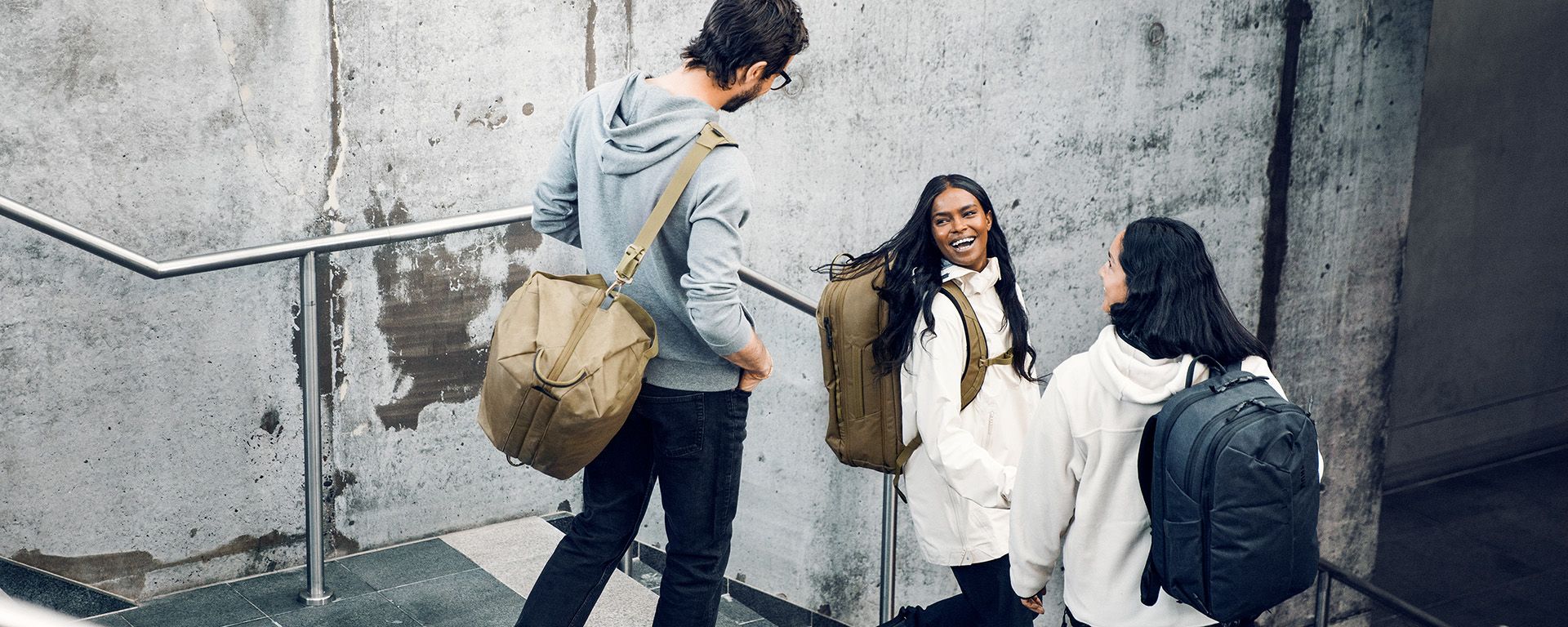 Three people walk down stairs with concrete walls laughing and carrying Thule Aion bags.