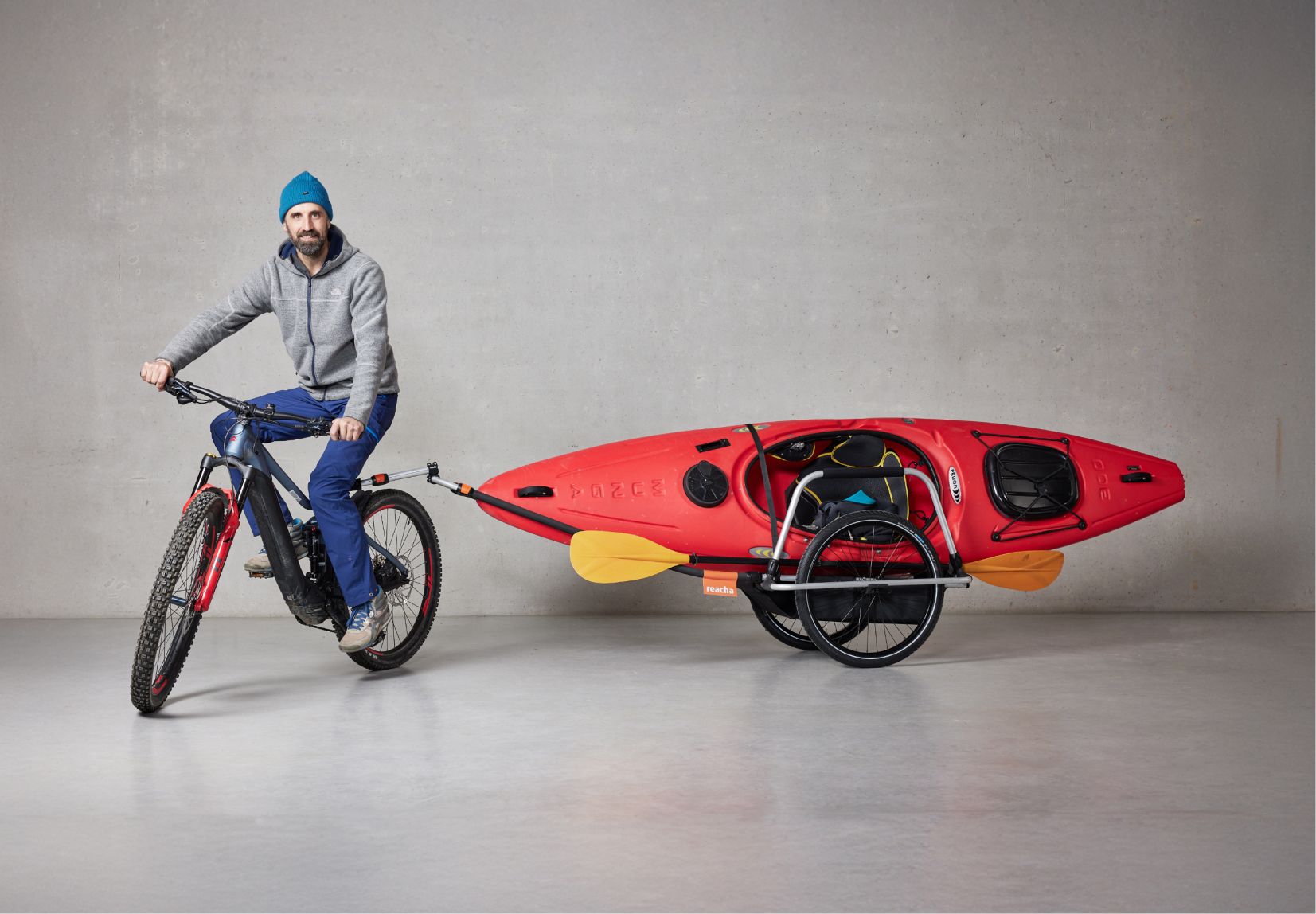Мужчина на стадионе едет на велосипеде с прицепом Reacha Sport, в который уложен ярко-оранжевый каяк.