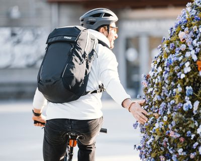 Un hombre anda en bicicleta llevando una mochila para bicicleta negra y acaricia flores mientras avanza.