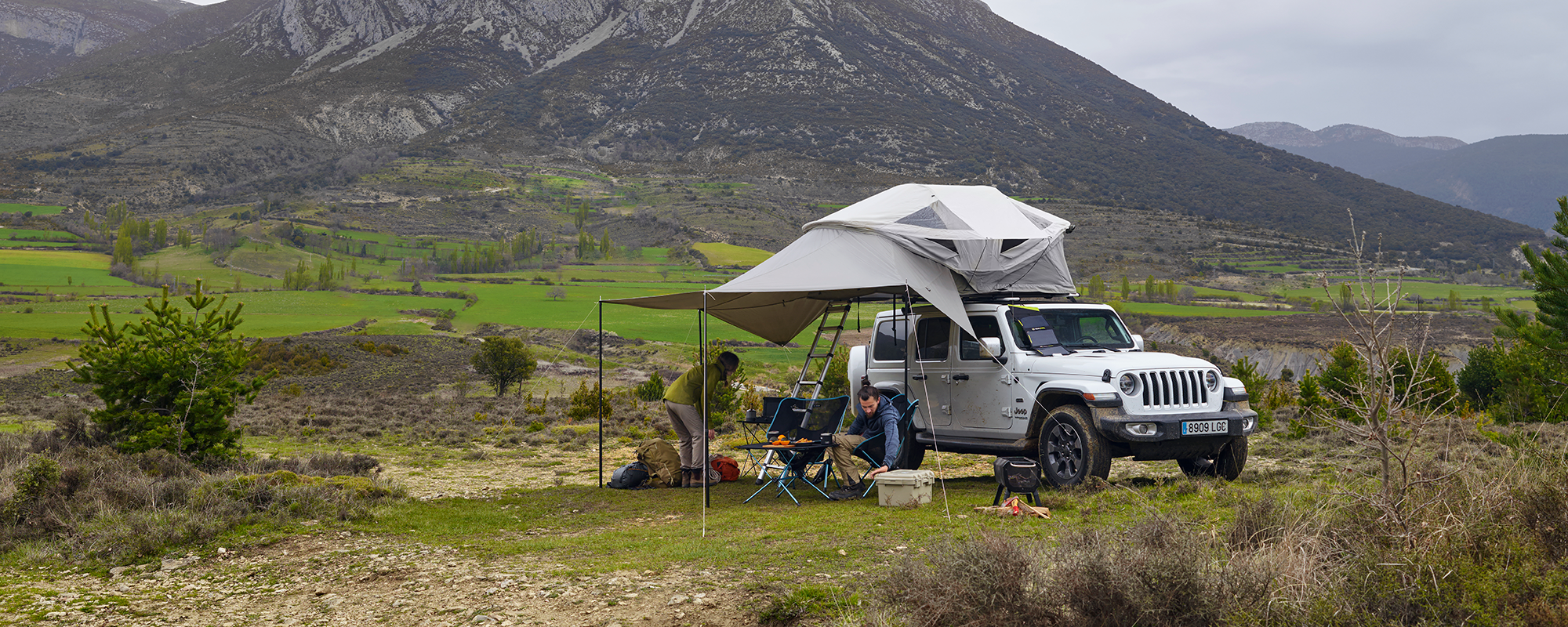 Two people sit in camping chairs under the awning of a jeep with a Thule Approach rooftop tent that is parked in the mountains.