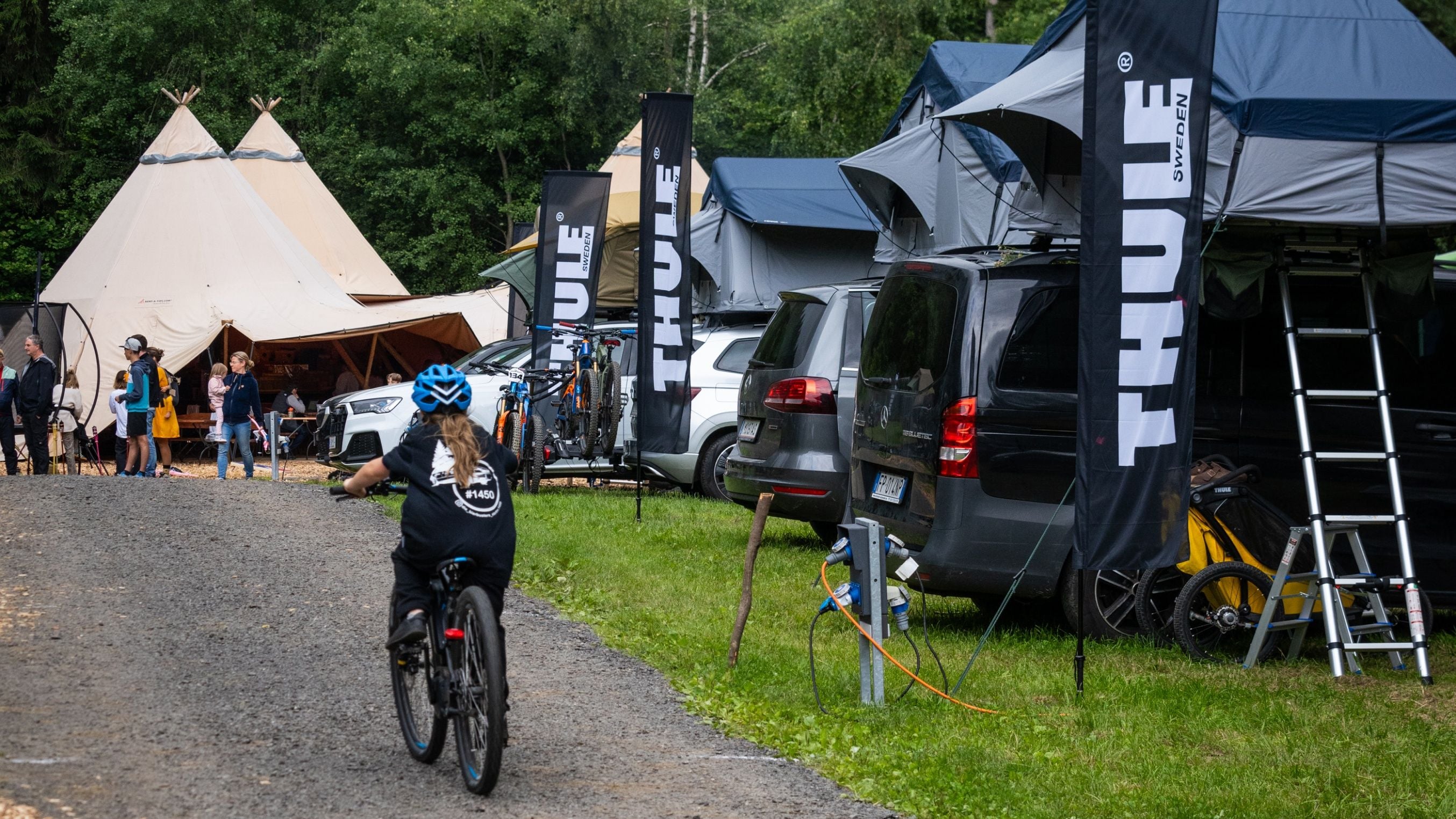 Niño andando en bicicleta en el evento Adventure Life by Thule en Innsbruck, con automóviles equipados con tiendas de techo Thule estacionados a un costado.