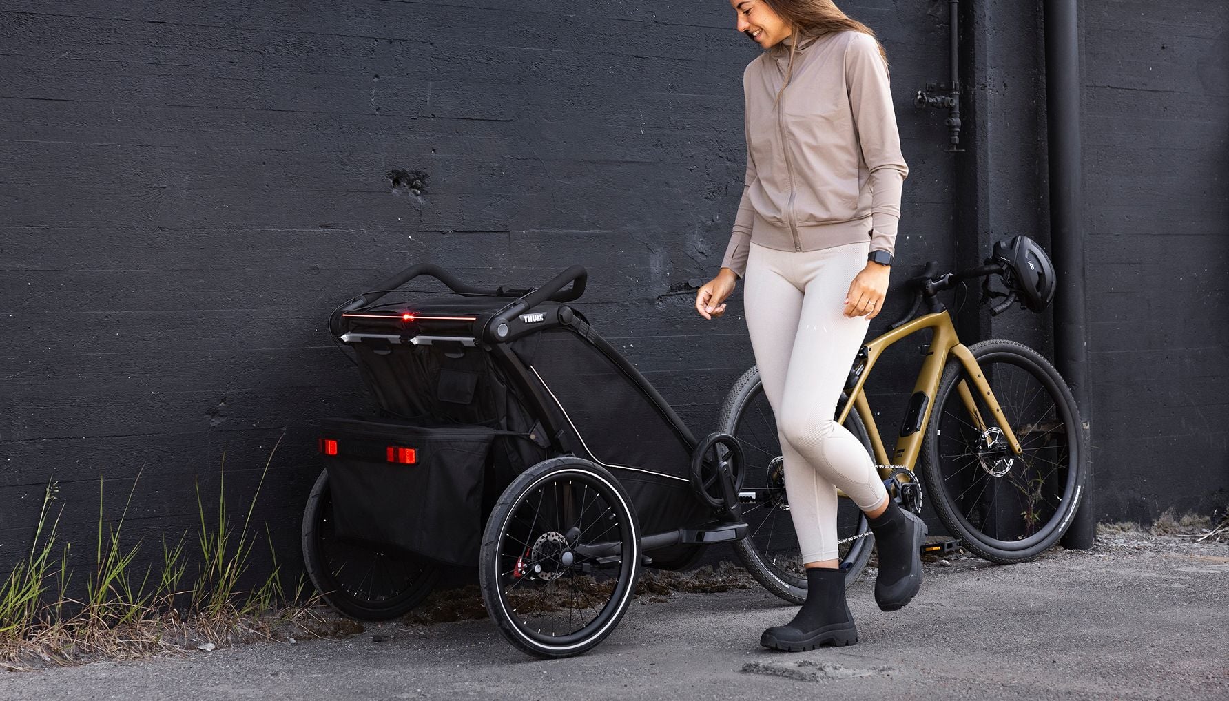 Person standing beside a bicycle with a bike trailer attached, next to a dark wall outdoors.