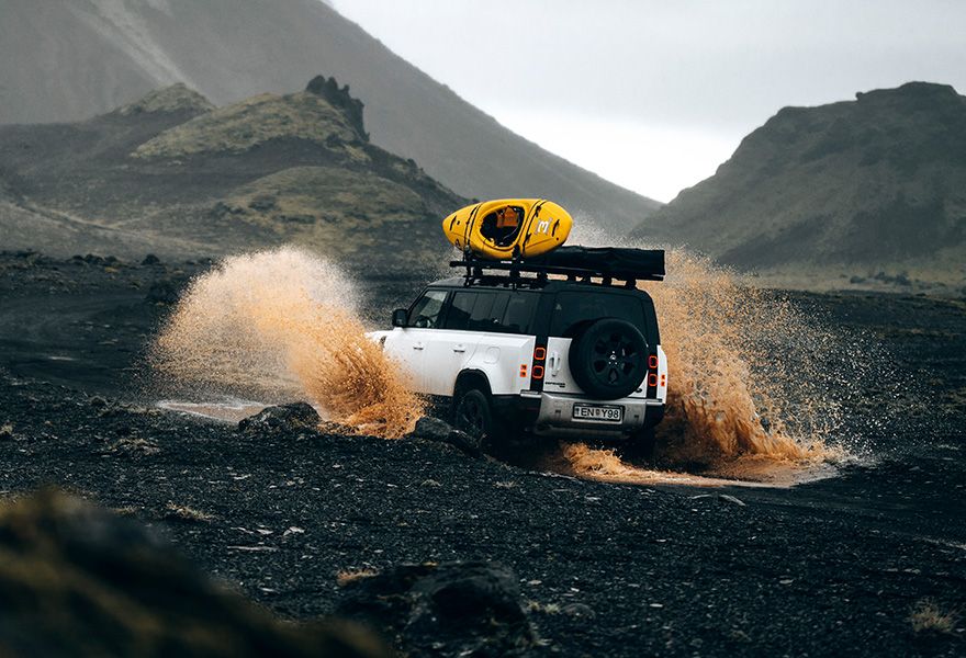 A car driving through water on a dirt road surrounded by mountains, carrying a kayak on a Thule Caprock roof platform. 