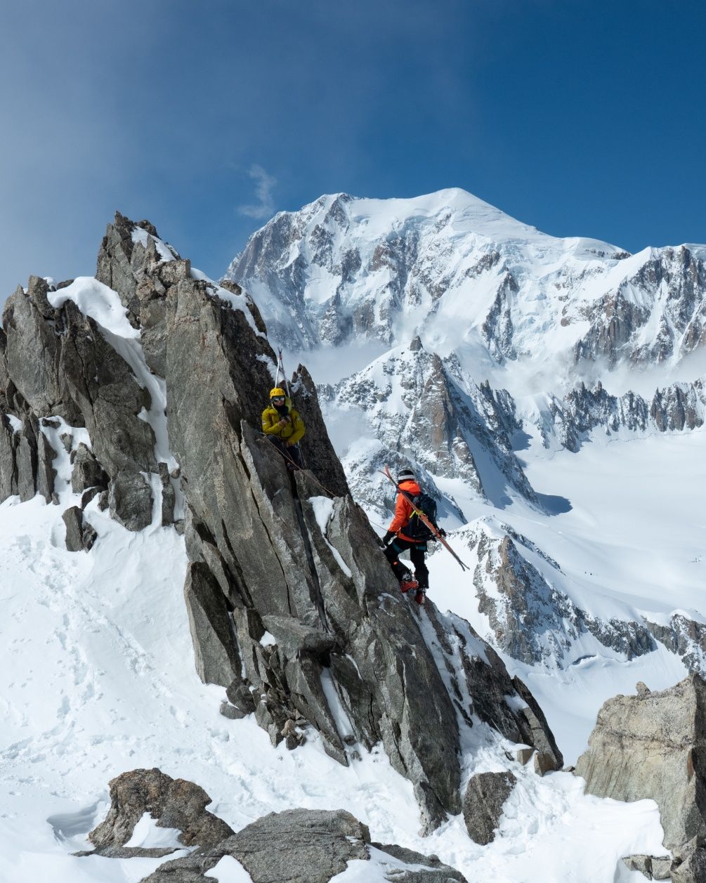 Two people with skis strapped to their backs climb a rocky mountain.