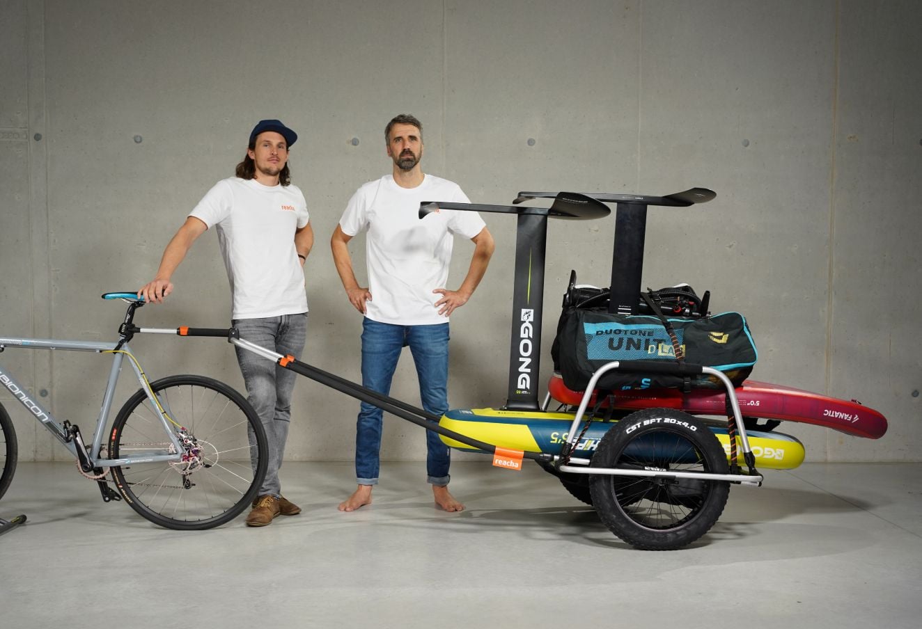 Två män poserar i en studio med en cykel fäst i en Reacha Sport Beach cykelvagn som bär surfbrädor och friluftsutrustning.