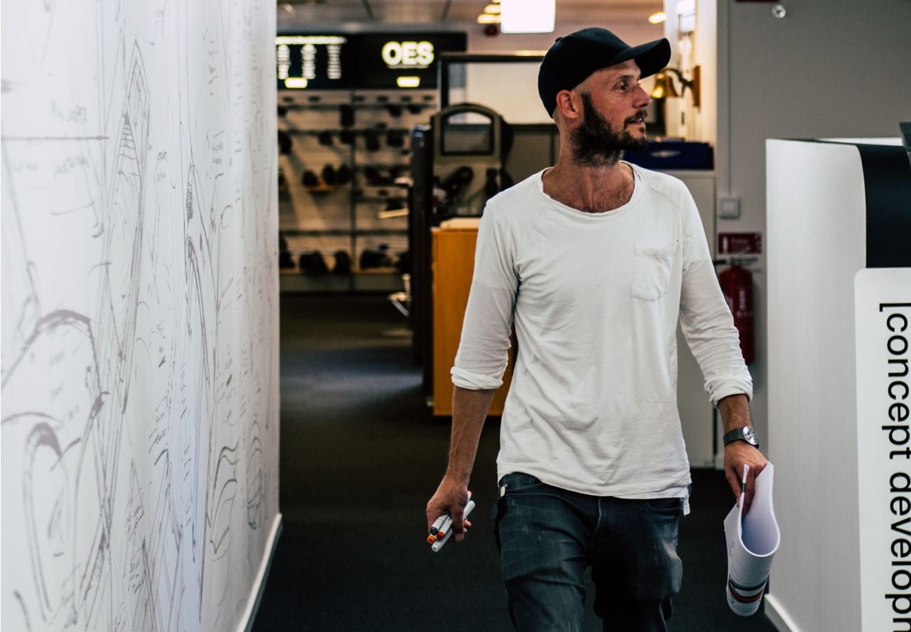 Un homme portant un t-shirt blanc, un jean bleu et une casquette noire marche dans un espace de bureau moderne tout en tenant une feuille roulée et un marqueur. Derrière lui, des croquis sont affichés aux murs et un environnement de travail est visible en arrière-plan.