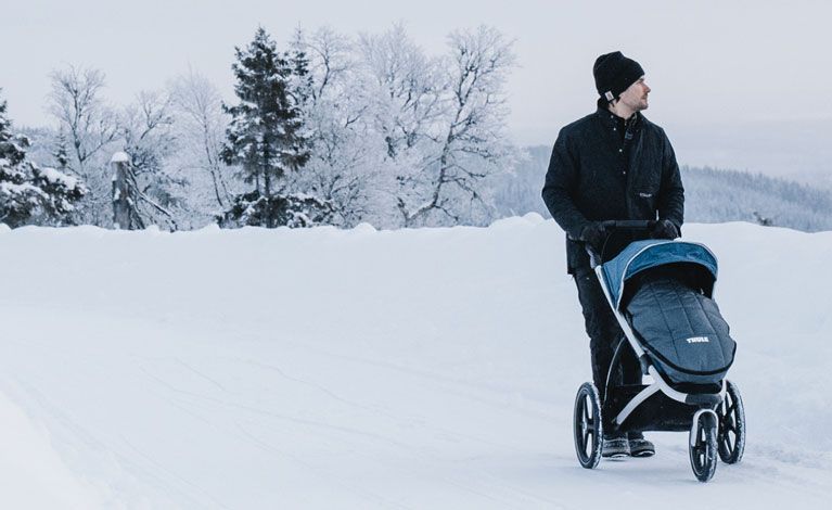A man walks through a snowy forest path with a the stroller Thule Urban Glide 2.