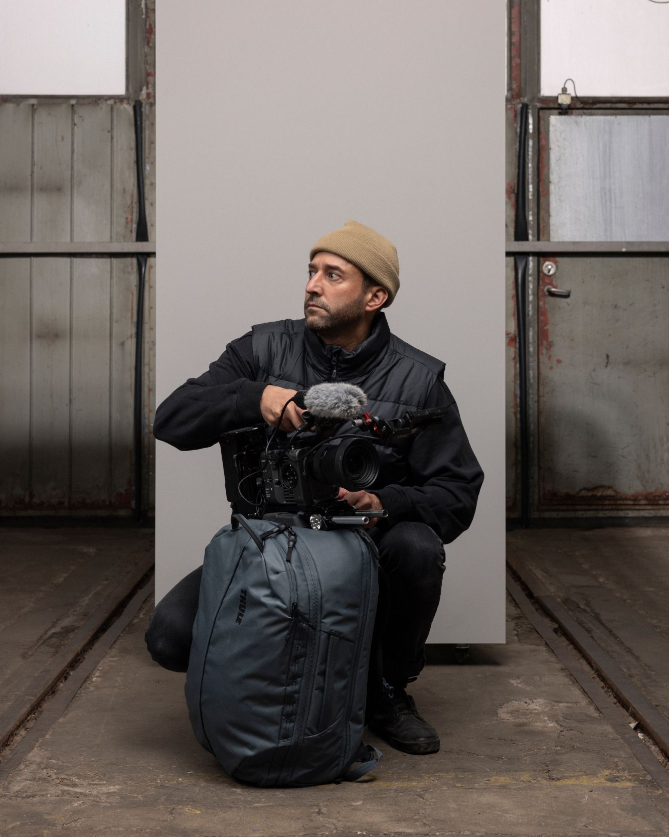 A man crouching down, looking to the side with a beige beanie and all black clothes. He has a Thule Aion bag in black in front of him and a big professional video camera with a microphone on top of it. 