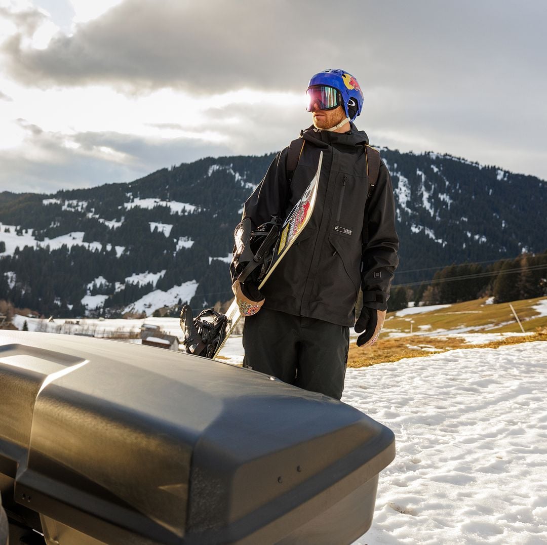 En snowboardåkare som står utomhus i vinterkläder, håller i en snowboard och tittar mot bergen.