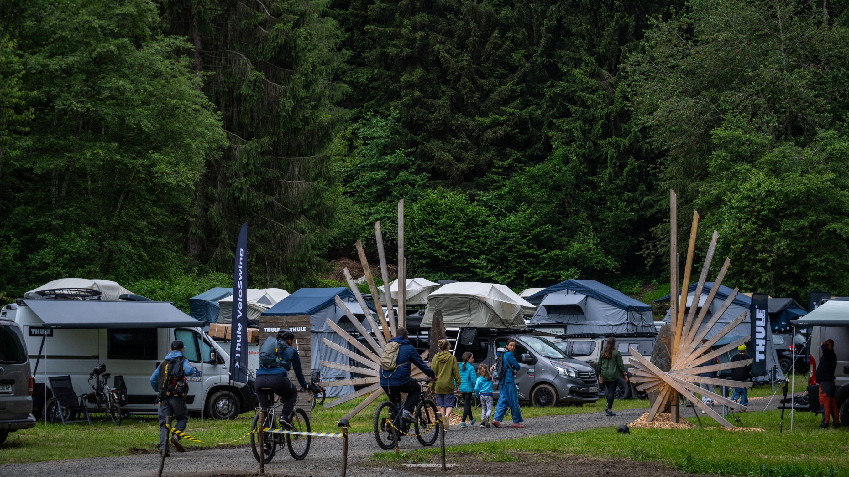 Участники прогуливаются и катаются на велосипедах по территории мероприятия Adventure Life by Thule, на фоне видны автомобили с палатками на крыше.