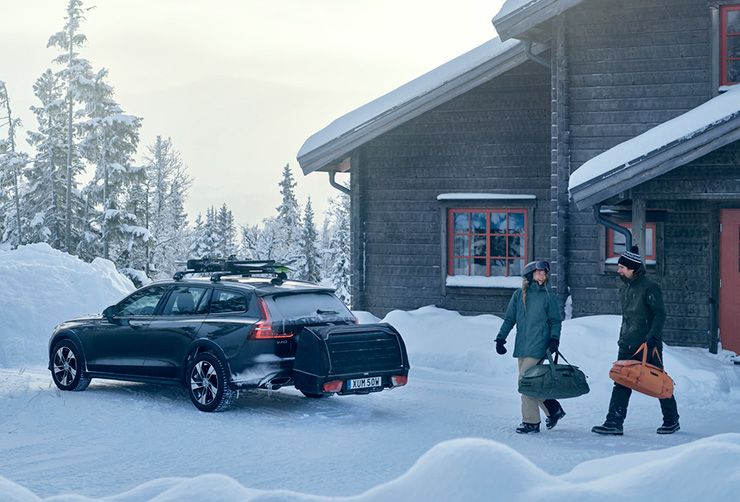Una pareja camina en la nieve hacia un vehículo eléctrico con un soporte para esquís en el techo y un portaequipajes trasero en el maletero.