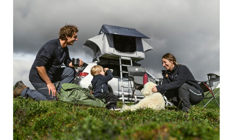 A family sit in the grass looking at their child with a Thule car top tent in the background.