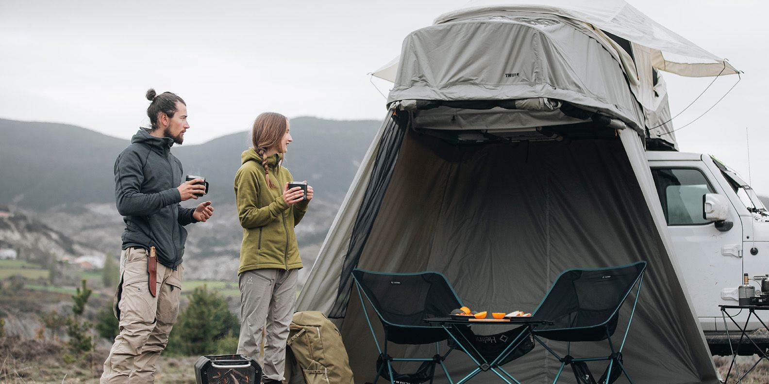 Due persone sono in piedi con bevande calde vicino alla loro tenda Thule Approach sul tetto dell'auto con una dépendance
