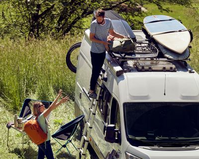 Un homme salue une femme qui charge son bagage dans un véhicule de loisir équipé de barres de toit chargées.