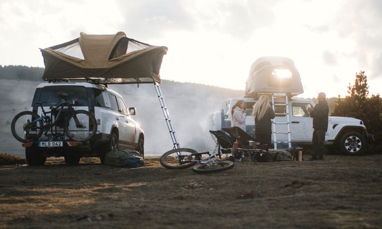Thuleルーフトップテントを積んだ2台の車が、人々がキャンプを設営している野原に駐車されています。