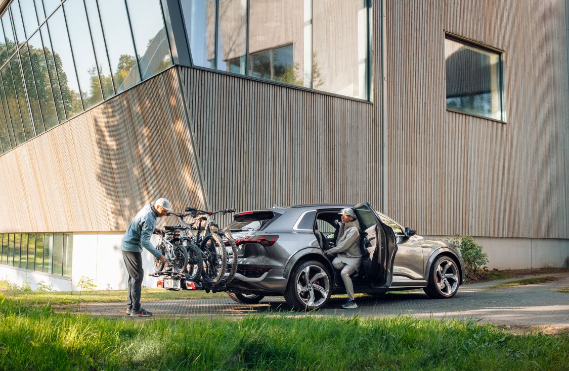 Hall auto, mille haakekonksule on paigaldatud jalgrattahoidik Thule EasyFold, on pargitud suure puumaja ette. Tagaistmel istub naine ja vaatab tagasi mehe poole, kes paneb jalgrattahoidikule nelja ratast.