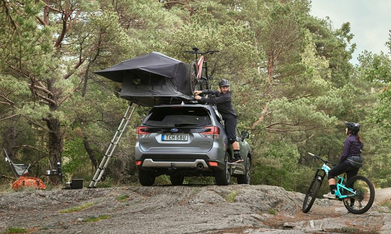 A cyclist unloads his bikes from a Thule roof bike rack where a Thule Tepui car top tent is also installed.