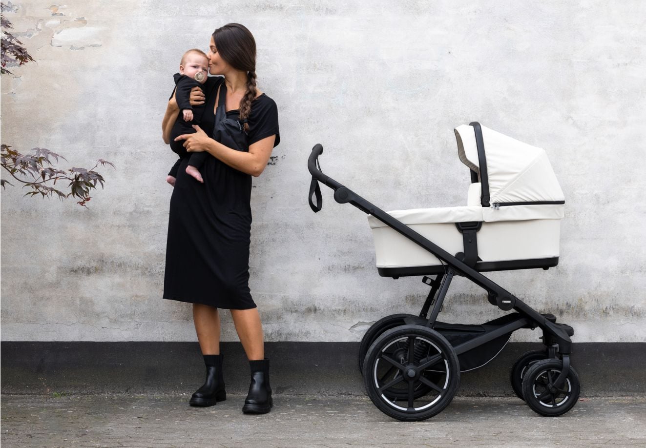 Une personne tient et embrasse un bébé tout en se tenant à côté d’une poussette à 4 roues Thule Urban Glide de couleur sable doux.