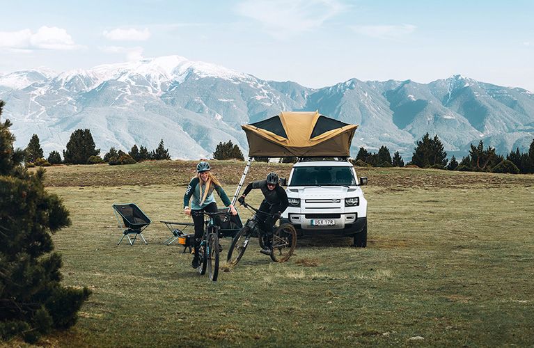 By the mountains, two cyclists cycle away in front of a car with a soft-shell rooftop tent.