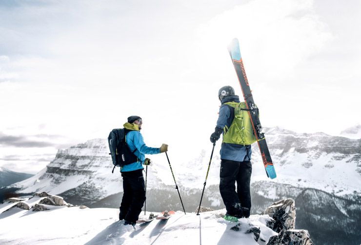 Two skiers stand on the top of a mountain with their Thule ski backpacks and ski gear.