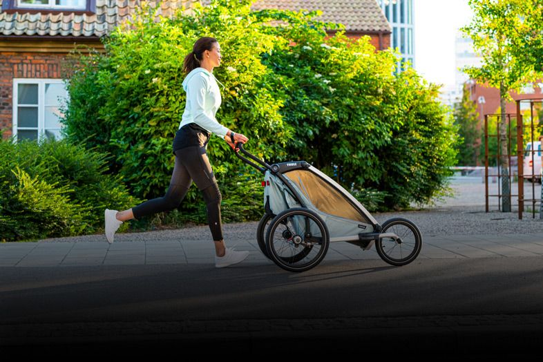 A woman in sportswear jogs down a residential street with a blue Thule Chariot Cross jogging bicycle trailer.