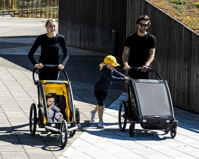 Двое родителей идут по залитой солнцем городской улице, катя велоприцепы для детей.