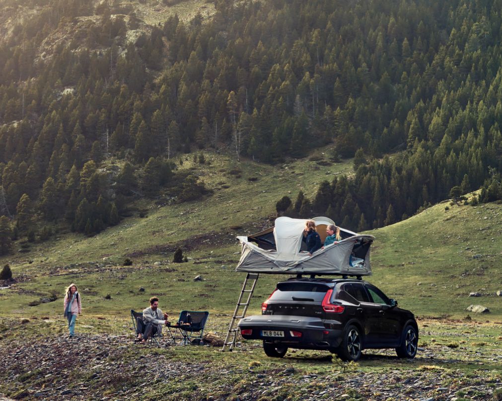 Un automóvil de color negro estacionado en un paisaje montañoso verde equipado con un portaequipaje para barra de remolque Thule Arcos y una tienda de techo Thule Approach. 