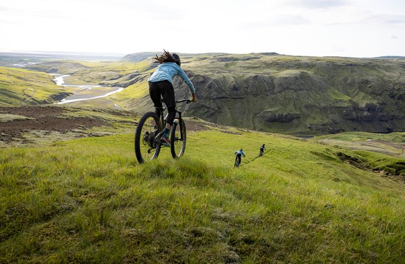 Un cycliste suit deux autres cyclistes au loin en descendant une colline verte sous un ciel bleu et une rivière.