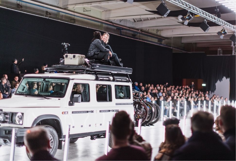 Two women sit on top of an SUV loaded with Thule gear while going down the runway at a fashion show.