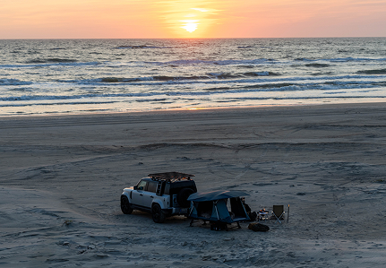 Um veículo está estacionado numa praia com uma tenda de reboque Thule Outset montada atrás. 