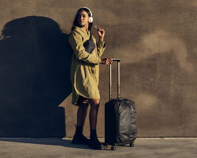 W słoneczny dzień kobieta stoi trzymając walizkę na kółkach.