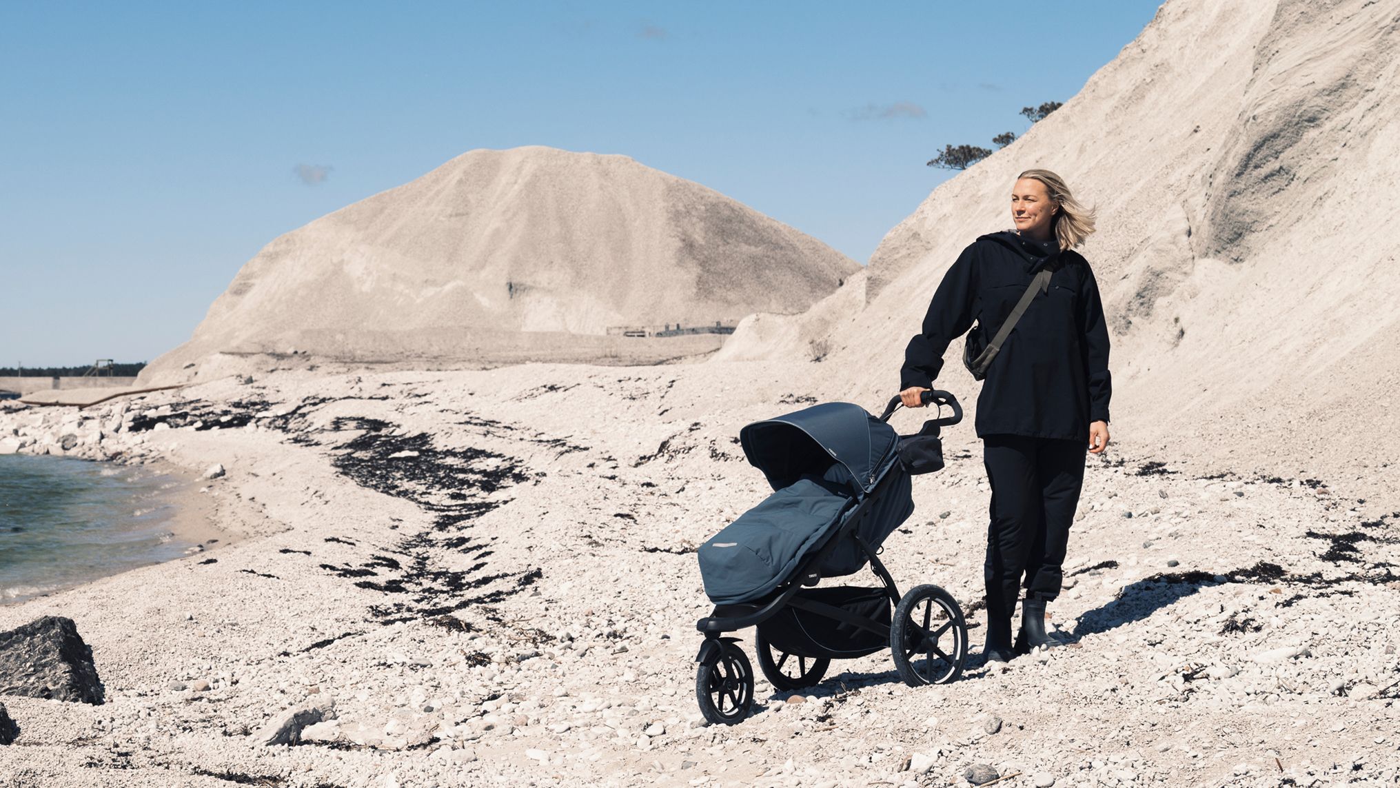 Sarah Sjöström ao lado de um carrinho Thule azul escuro em uma costa rochosa, cercada por penhascos de calcário pálido sob um céu azul claro.