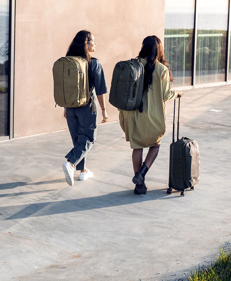 Two women walk down a sunny sidewalk with Thule Aion backpacks and carry on suitcase.