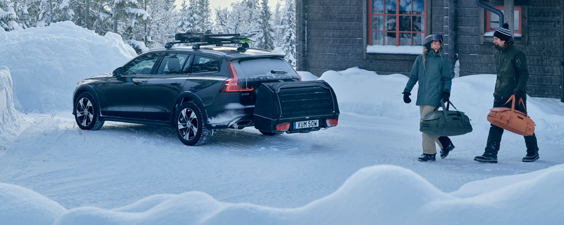 A car with a Thule Onto rear cargo carrier is parked in the snow by a cottage with a ski rack.