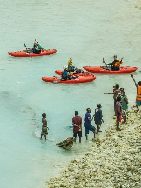 A zoomed out image of people in 4 red kayaks waving to people on the shore.
