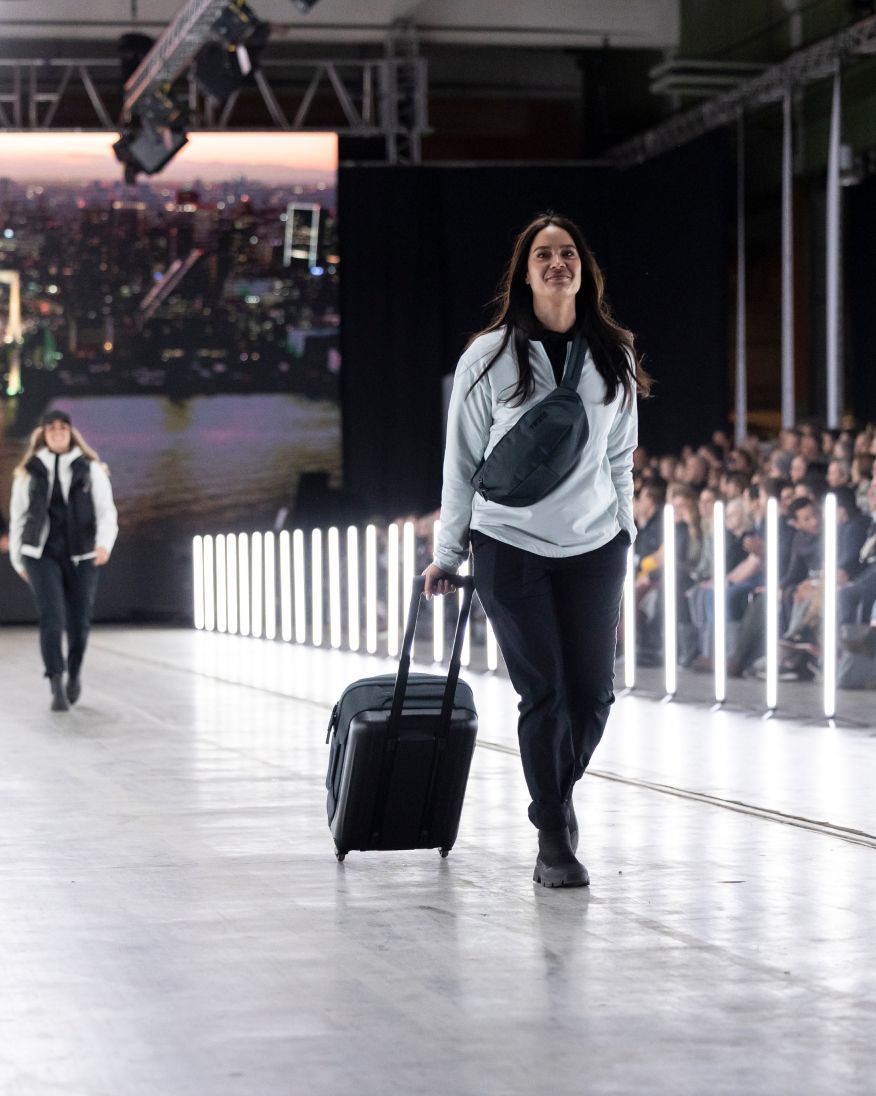 A woman with long brown hair walking down a fashion runway with a Thule Subterra 2 cross-body bag and rolling suitcase. Behind her is another woman walking down the runway and on the side is a crowd of people looking at them.