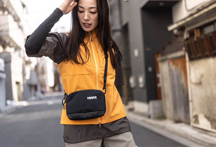 A woman stands on a city street with an orange jacket and a black Thule Paramount sling bag.