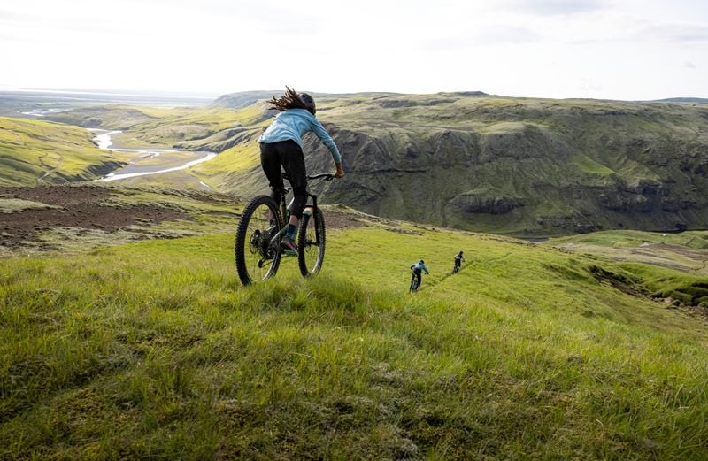 En cyklist følger to andre cyklister i det fjerne ned ad en grøn grasbakke med blå himmel og en flod.