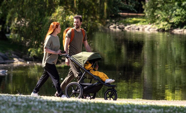 By a pond, a couple talk while the woman pushes a child in a Thule stroller.