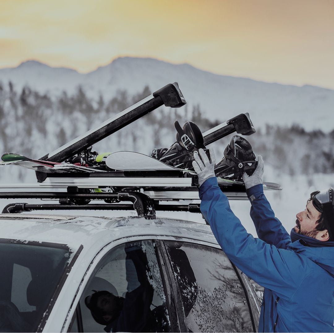 Person loading skis onto a Thule roof platform on a car in a snowy mountain landscape. 